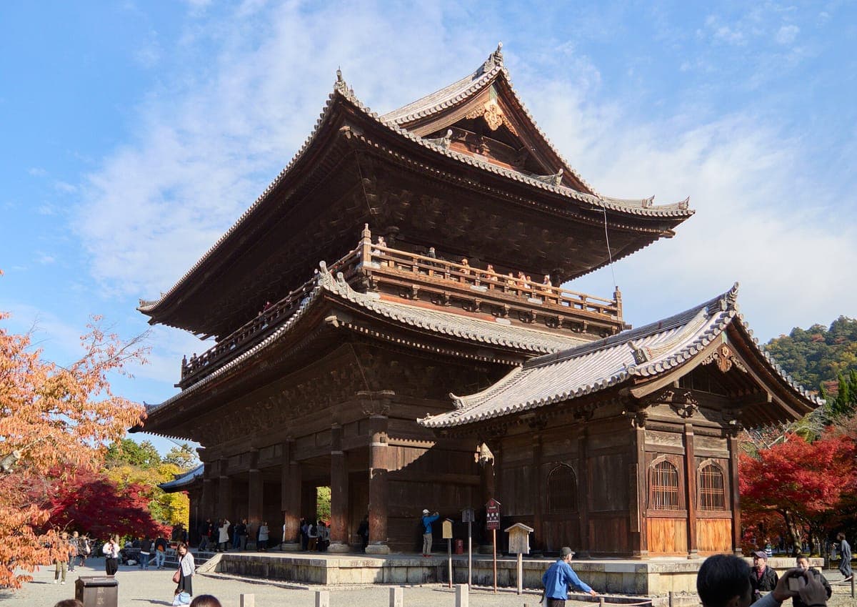 L'immense porte en bois Sanmon à deux étages du Nanzen-ji sur fond de ciel bleu, avec des érables rouges d'automne de chaque côté et des visiteurs à sa base