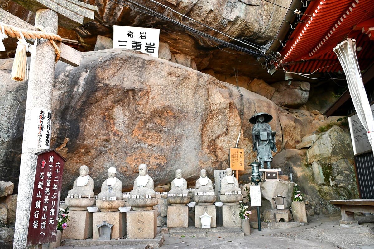 Řada malých kamenných soch Buddhy před obrovským balvanem v Senkō-ji chrámu v Onomichi, s kamennou torii vlevo a červenou budovou chrámu vpravo