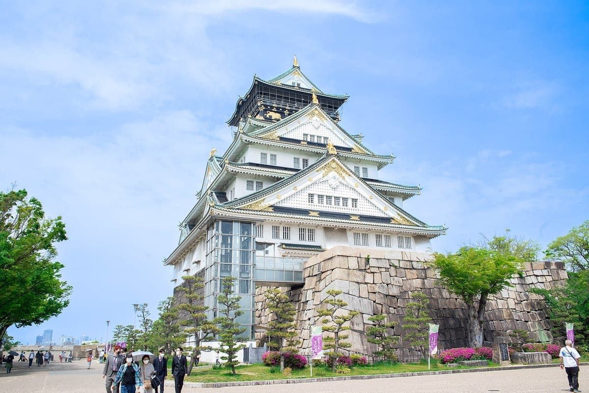 La torre principal blanca del Castillo de Osaka con techos verdes y decoraciones doradas, elevándose sobre un alto muro de piedra bajo un cielo azul
