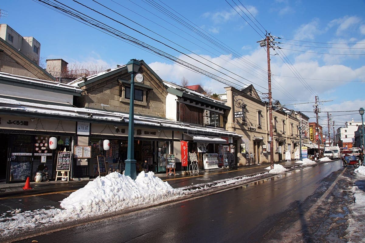 Sakaimachi-dōri v Otaru, ulice nízkých starých obchodních budov s taškovou střechou a nápisy obchodů, nahromaděný sníh podél obrubníku a mokrá silnice