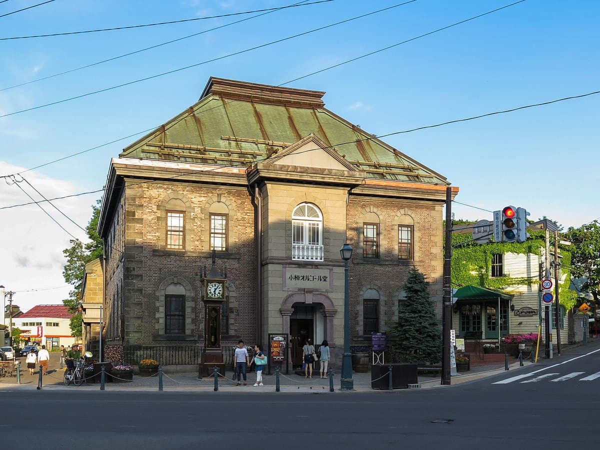 Dvoupatrová kamenná a cihlová hlavní budova Otaru Music Box Museum se zelenou měděnou střechou, na rohu ulice pod modrou oblohou