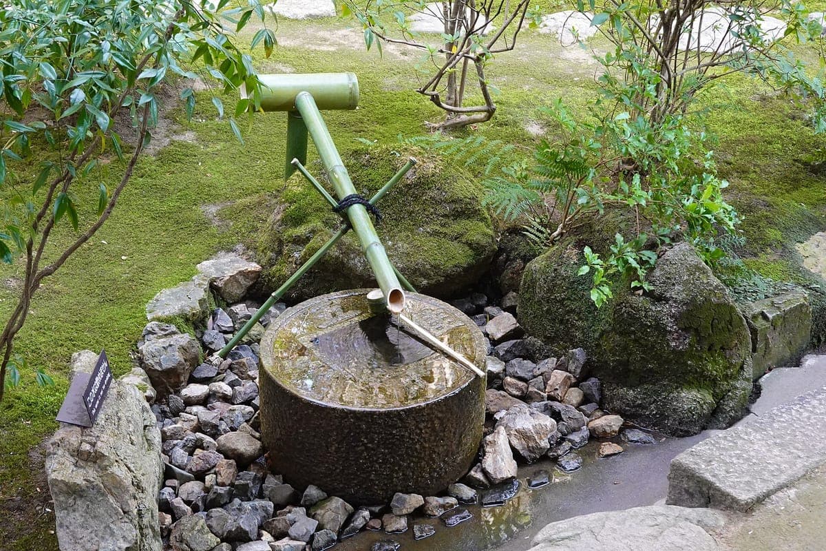La palangana de piedra redonda tsukubai en Ryōan-ji con un agujero cuadrado en el medio, una tubería de bambú que gotea agua y un cucharón, rodeada de piedras y musgo.