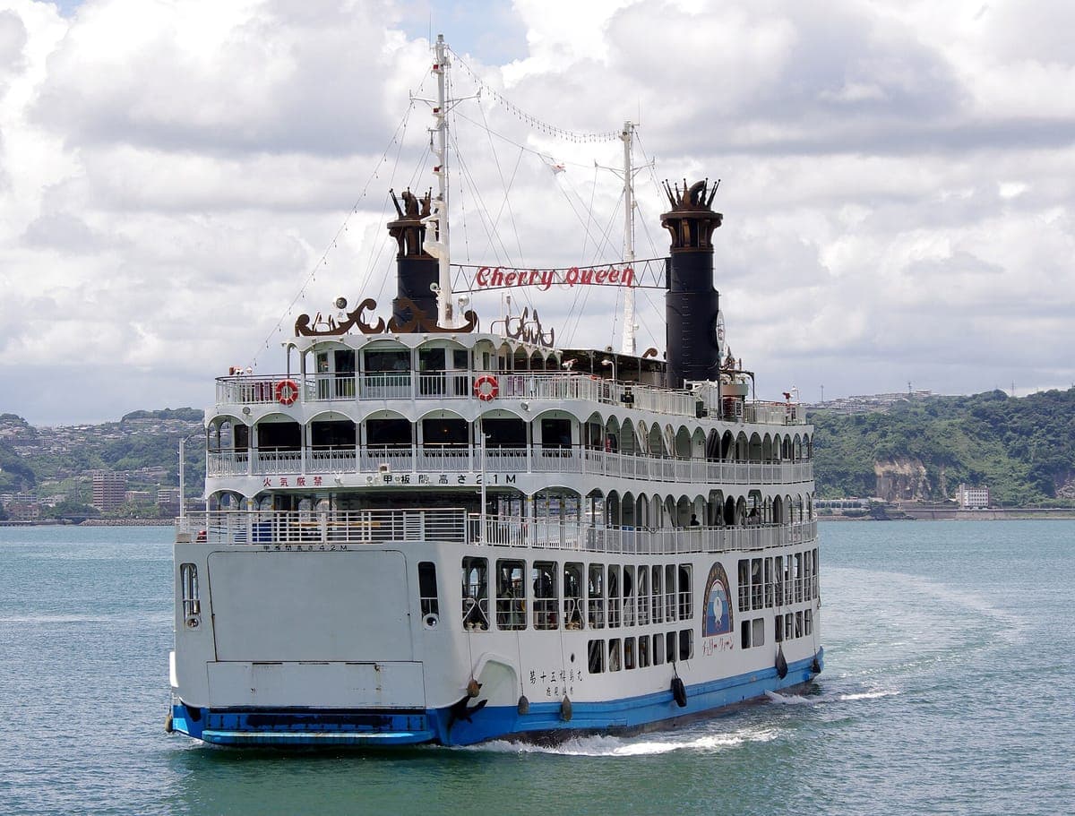 Isang puting multi-deck na Sakurajima ferry na may dalawang itim na funnel na tumatawid sa tahimik na tubig ng Kagoshima Bay, kasama ang baybayin ng lungsod sa likod