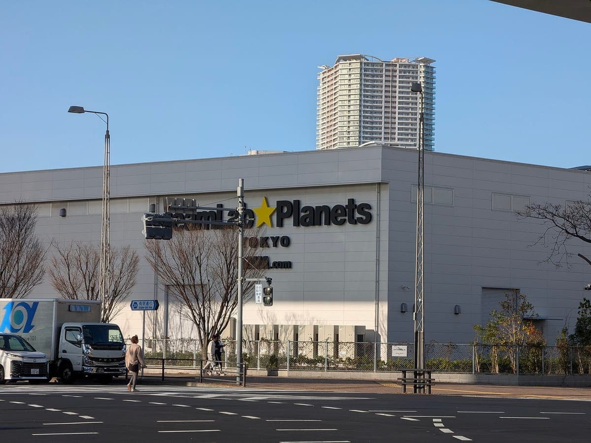 Šedý exteriér teamLab Planets TOKYO připomínající skladiště se svým jménem na zdi, širokým přejezdem silnice a věžovým blokem za ním