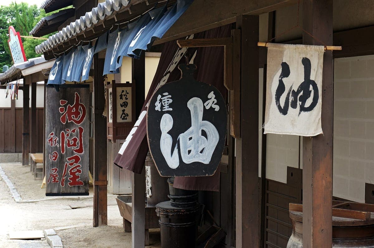 Isang Edo-style na kalye sa film village na may mga karatula sa tindahan at mga kurtina ng noren, kabilang ang isang malaking itim na signboard na may kanji para sa langis at isang puting banner