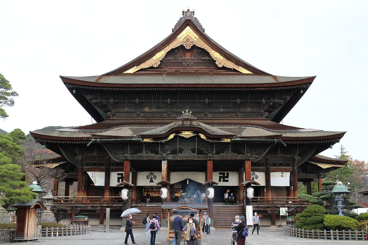 Před Zenkō-ji's hlavní halou v Naganu, obrovská tmavá dřevěná budova s dvojitou střechou, zlaceným štítem a kadidelnicí vpředu, s návštěvníky na nádvoří