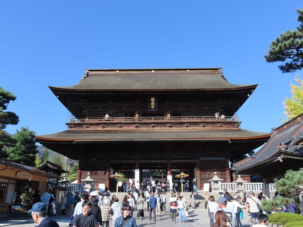 Dvoupatrová dřevěná Sanmon brána Zenkō-ji pod modrou oblohou, s kamennou dlažbou a návštěvníky, kteří k ní jdou